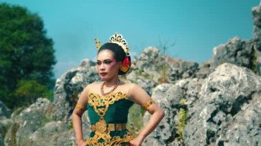 Bir kadın, kayalık bir arazide yeşil ve altın rengi bir kostümle geleneksel bir dans sergiliyor. Özenle hazırlanmış makyaj ve aksesuarları var, sakin bir ifadesi var, kültürel mirası ve sanatını sergiliyor..