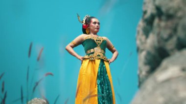 Genç bir kadın, karmaşık altın süslemeleri olan yeşil ve sarı bir kıyafetin yer aldığı geleneksel kıyafetlerle güven içinde poz veriyor. Uzun çimenler ve kayalık oluşumlarla çevrili parlak mavi bir gökyüzüne karşı duruyor. Zarafet ve güç saçıyor..