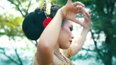 Canlı makyaj ve dekoratif başlıkla süslenmiş geleneksel bir dans sergileyen bir kadının yakın çekimi. Saçında çiçekler var ve ellerini zarif bir şekilde hareket ettiriyor, bulanık doğal bir arka plana doğru..