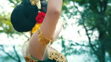 Geleneksel bir dans sergileyen, canlı makyaj yapan ve renkli bir kostüm giyen bir kadın. Ağaçlarla çevrili doğal bir arka plan üzerine kurulu anlamlı bir jestle elleriyle poz veriyor..