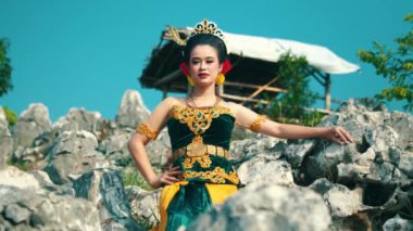 Geleneksel giyinmiş bir kadın, açık hava ortamında güven içinde poz verir. Karmaşık desenleri olan, taç ve kırmızı aksesuarlarla tamamlanmış güzel tasarlanmış siyah ve altın bir elbise giyiyor.