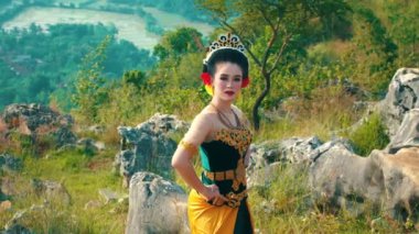 Geleneksel giyinmiş bir kadın, açık hava ortamında güven içinde poz verir. Karmaşık desenleri olan, taç ve kırmızı aksesuarlarla tamamlanmış güzel tasarlanmış siyah ve altın bir elbise giyiyor.