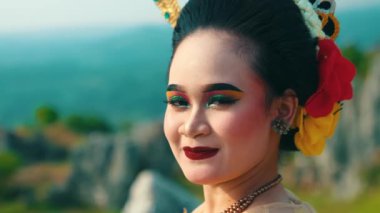 Renkli göz farı ve kalın rujlu canlı makyajlı bir kadının profil portresi. Saçında geleneksel aksesuarlar var, çiçekler de dahil. Arka plan bulanık bir manzara. 