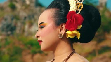 Saçlarında renkli çiçeklerle süslenmiş geleneksel makyaj ve giysili genç bir kadın, doğal bir arkaplana karşı güvenle ayakta duruyor. Yüzü dingin, göz makyajı ve kalın bir ruj..