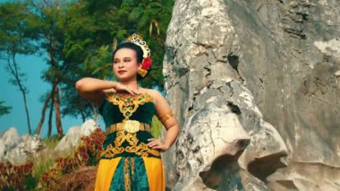 Geleneksel giyinmiş bir kadın, kayalık bir zemine güvenle poz verir. Parlak yeşil ve sarı bir elbise giyiyor. Saçında karmaşık altın ayrıntıları ve çiçek aksesuarlarıyla süslenmiş. Kültürel mirası gösteriyor..