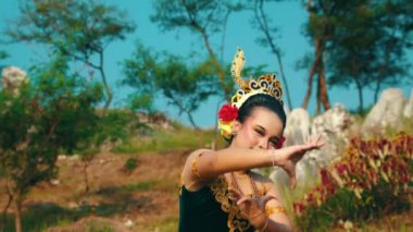 Altın aksanıyla süslenmiş canlı yeşil bir kostümle geleneksel dans eden bir kadın. Etrafı ağaçlar ve kayalarla çevrili doğal bir manzarayla çevrili. Kültürel mirası ve sanatını sergiliyor..