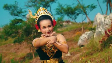 Doğal bir ortamda geleneksel bir dans gösterisi yapan bir dansçı. Dansçı karmaşık tasarımları olan renkli bir kostüm giyiyor, kültürel mirası gözler önüne seriyor. Arkaplanda yeşil ve kayalık arazi var, canlı atmosferi geliştiriyor..