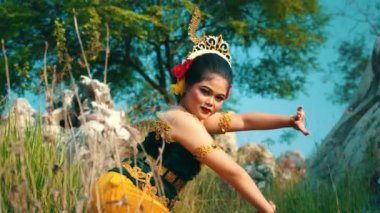 Genç bir kadın yemyeşil bir açık hava ortamında geleneksel bir dans sergiliyor. Karmaşık aksesuarları olan renkli bir kostüm giyiyor. Kültürel mirası gözler önüne seriyor. Arka planda ağaçlar ve kayalar var, canlı atmosferi geliştiriyorlar..