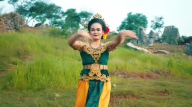 Bir kadın yemyeşil bir manzarada geleneksel bir dans sergiliyor. Renkli bir kostüm giyiyor. Karmaşık desenler ve aksesuarlarla süslenmiş. Arka planda kayalık oluşumlar ve ağaçlar yer alıyor, canlı bir kültürel manzara yaratıyor..