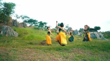 Bir grup dansçı yemyeşil bir arazide geleneksel dans gösterisi yapıyorlar. Canlı sarı ve yeşil kostümler giyiyorlar. Kayalık oluşumlar ve ağaçlar karşısında kültürel mirası sergiliyorlar..