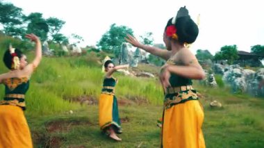 Geleneksel Endonezya kıyafeti giymiş beş kadından oluşan bir grup, kırsal bir ortamda dans ediyorlar. Renkli batik giysiler giyip kuşaklar takıyorlar. Kültür mirası sergiliyorlar..