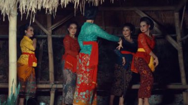 Bir grup kadın renkli kostümlerle geleneksel dans gösterisi yapıyorlar. Sahnede sazdan çatılı bir bambu yapısı yer alıyor. Dansçılar, çiçek desenleri ve kuşaklarla süslenmiş, karmaşık hareketler sergiliyorlar..