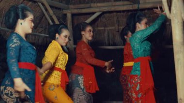 Renkli giysiler içinde geleneksel bir dans sergileyen bir grup kadın. Karmaşık kostümler ve zarif hareketler sergiliyorlar. Arka planda bambu yapıları olan kültürel bir mekan gibi görünüyor..