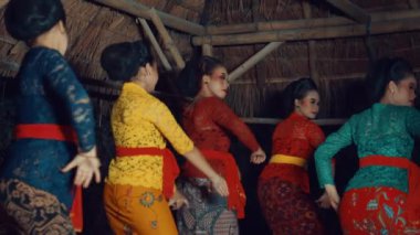 Geleneksel Endonezya kıyafeti giymiş beş kadından oluşan bir grup, kırsal bir ortamda dans ediyorlar. Renkli batik giysiler giyip kuşaklar takıyorlar. Kültür mirası sergiliyorlar..