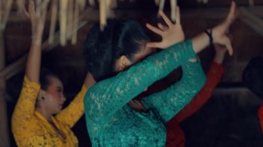 Bir grup dansçı renkli giysiler içinde geleneksel dans gösterisi yapıyorlar. Ön plandaki ana dansçı kırmızı kuşaklı yeşil dantel bir elbise giyerken, arka plandaki diğer dansçılar sarı ve kırmızı elbiseler giyer.. 