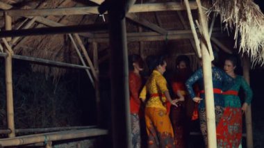 Bambu bir yapının içinde geleneksel bir dans sergileyen bir grup kadın. Renkli kostümler giyiyorlar. Karmaşık desenleri var. Kültürel mirası sergiliyorlar. Ayarlar loş, hareketlerini ve ifadelerini vurguluyor.