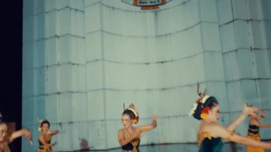 Renkli kostümlü dansçıların olduğu geleneksel bir dans gösterisi. Tepesinde bir amblem olan büyük, dokulu bir duvara karşı yapılmış. Dansçılar hareket halinde, karmaşık el hareketleri ve ifadeleri sergiliyorlar, kültürel mirası somutlaştırıyorlar..