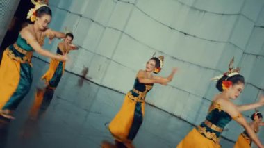 Geleneksel Endonezya kıyafeti giymiş beş kadından oluşan bir grup, kırsal bir ortamda dans ediyorlar. Renkli batik giysiler giyip kuşaklar takıyorlar. Kültür mirası sergiliyorlar..