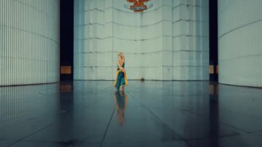 Modern mimari arkaplana karşı canlı bir kostüm içinde zarif bir dansçı. Sahne, dansçının hareketlerini ve etrafındaki parlak yüzeyleri aydınlatıyor..