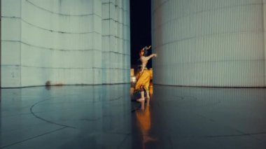 Çarpıcı sarı ve siyah bir kıyafetle dans eden bir dansçı modern mimari alanda geleneksel hareketler sergiliyor. Arka planda, dansçının dinamik duruşunu geliştiren pürüzsüz, yansıtıcı bir zemine sahip geniş, kıvrımlı duvarlar yer alıyor..