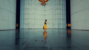 Canlı sarı elbiseli bir dansçı geniş silindirik yapıları olan endüstriyel bir ortamda gösteri yapar. Yumuşak, yansıtıcı zemin, yumuşak ışıkla aydınlatılan dramatik atmosferi güçlendirir..