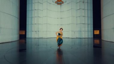 Bir dansçı modern mimari alanda zarif bir performans sergiliyor, uzun, silindirik yapıları var. Zemin dansçının hareketlerini yansıtıyor, dinamik bir görsel etki yaratıyor. Işıklandırma dramatik, sahnenin atmosferini arttırıyor..