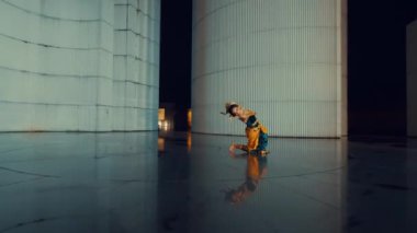 Modern mimari alanda zarif bir performans sergileyen bir dansçı. Arka planda geniş silindirik yapılar var. Dansçı renkli bir kıyafet giyer. Yansıması parlak zeminden görülebilir. Dinamik bir görsel efekt yaratır..