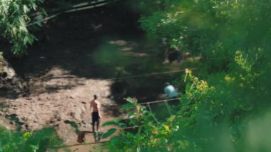 A group of people working in a muddy area near a stream, surrounded by lush greenery. Some individuals are partially clothed, engaged in activities related to the water or mud.