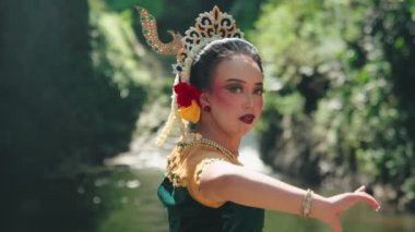 Geleneksel kıyafetli bir dansçı, bereketli yeşilliklerle çevrili sakin bir nehirde zarifçe dans eder. Dansçının makyajı canlı, etkileyici özelliklerini vurguluyor ve süslü bir takı ve dekoratif bir başlık takıyor..