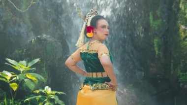 Geleneksel giyinmiş bir kadın, yemyeşil bir şelalenin önünde zarifçe poz verir. Kıyafetinde karmaşık tasarımlar ve canlı renkler var. Kültürel mirası vurguluyor..