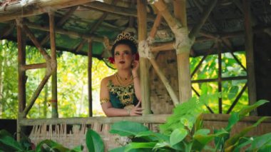 Geleneksel Bali kıyafetli bir kadın, yemyeşil bir bambu yapının içinde zarifçe poz verir. Makyajı çok canlı ve kültür mirasını yansıtan süslü mücevherler takıyor..