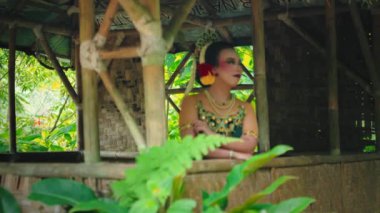Geleneksel giyinmiş bir kadın, bereketli yeşilliklerle çevrili bambu bir yapıda zarifçe poz verir. Karmaşık takılar takıyor ve saçına çiçekli bir aksesuar takıyor. Zarafet ve kültürel güzellik yayıyor..