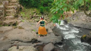 Geleneksel giyinmiş bir kadın, yemyeşil sularla çevrili, akan bir derenin kenarında kayaların üzerinde durur. Dans ediyor ya da kültürel bir ayin yapıyor gibi görünüyor. Arka planda dingin bir şelale var..
