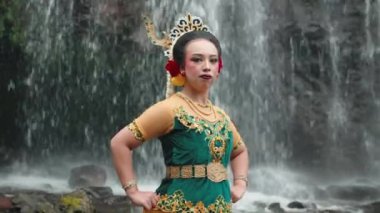 Geleneksel Endonezya kıyafeti giymiş genç bir kadın şelalenin önünde güven içinde poz veriyor. Saçındaki çiçek aksesuarlarıyla tamamlanmış, karmaşık desenlerle süslenmiş yeşil ve altın bir elbise giyiyor..