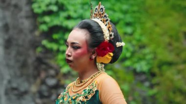 Renkli bir kostüm giymiş geleneksel bir dansçı, çiçeklerle süslenmiş ve karmaşık mücevherlerle süslenmiş, yemyeşil bir arka planda zarif bir gösteri sergiliyor..