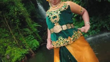 Geleneksel giyinmiş bir kadın yemyeşil bir nehir kıyısının yanında poz veriyor. Karmaşık nakış ve canlı renkler sergiliyor. Arka planda yoğun yeşillik ve akan su, sakin bir atmosfer yaratıyor..