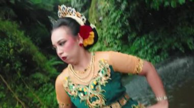 Zengin yeşil ve altın kumaşlı, karmaşık nakış ve süslemelerle süslenmiş geleneksel bir kıyafetin yakın çekimi. Dekor, canlı kıyafete dingin bir zemin ekleyerek, bir su kütlesinin yakınında gibi görünüyor..