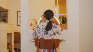 Saç bakım seansını bitiren bir kadının arka açısı. Dokusunu kontrol etmek için ellerini yeni taranmış saçlarında gezdiriyor. Dairesinde rahat bir şekilde oturuyor. Yuvarlak aynaya bakıyor..