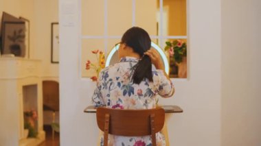 Aydınlatılmış yuvarlak aynanın önünde oturan uzun siyah saçlı genç bir kadının arka görüntüsü. O, sıcak bir yatak odası atmosferinde akşam güzellik rejimine başlamak için saç bağını çözüyor..