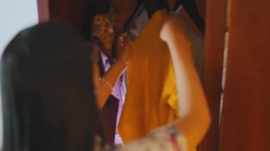 Elbise seçmek için ahşap bir dolaba uzanan genç bir kadının arka görüntüsü. Günlük giysisine sarı bir elbise seçmek için askıları karıştırıyor..