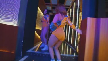 Üç dansçının, sarı şort ve ceket giymiş lüks bir koridorda kameradan uzaklaşmalarını gösteren bir arka görüntü..