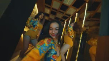 Modern bir iç mekanda merdivenlerde poz veren üç genç kadın, kulübün içinde sarı desenli kıyafetler giyiyorlar.