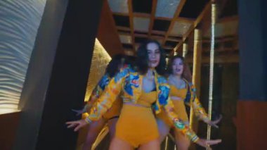 Üç dansçı lüks bir koridorda ritimli bir şekilde poz veriyor ve hareket ediyorlar. Sarı şehir kıyafetleri içinde güven ve tarz gösteriyorlar..