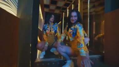 Üç dansçı aynalı bir koridorda çömelip poz vererek kameraya sert bir ifadeyle bakıyor..
