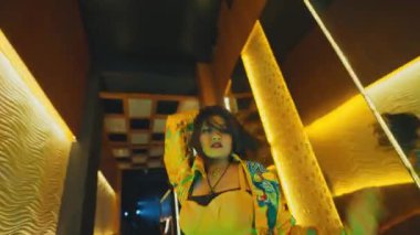 Aynalı bir koridorda devriye gezen bir kadın dansçının yakın görüntüsü. Saçlarını savuruyor ve lüks bir gece kulübünde dans ederken tavırlarını sergiliyor..