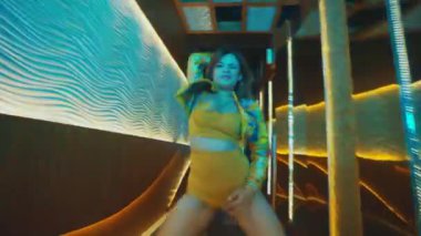 Bir bayan dansçı, sarı iki parçalı elbisesiyle lüks bir koridorda dans ederken dramatik bir saç taklası yapar..