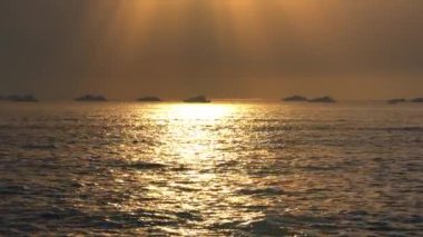 Gün batımında sakin bir deniz manzarası, sakin sulara yansıyan altın güneş ışığı. Uzak teknelerin siluetleri ufukta görünüyor ve sakin bir atmosfer yaratıyor..