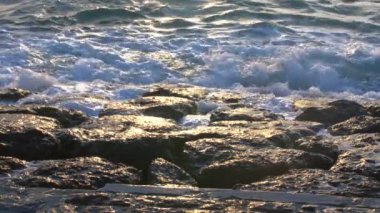 Kayalık kıyı şeridinin sakin bir görüntüsü. Dalgalar yavaşça kayalara çarpıyor. Güneş ışığını yansıtıyor. Su parlıyor, sakin bir atmosfer yaratıyor..