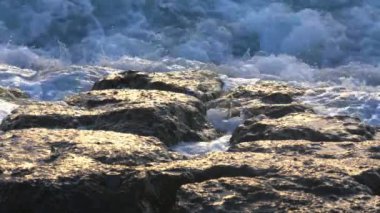 Dalgalar kayalık sahile çarpıyor, köpük ve su sıçratıyor. Güneş ışığı ıslak kayalardan yansıyarak, sahnenin dokusunu ve rengini güçlendirir..