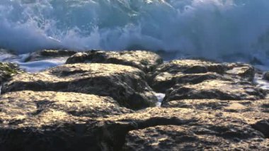 Dalgalar kayalık sahile çarpıyor, köpük ve su sıçratıyor. Güneş ıslak kayalardan yansıyarak, dokularını ve renklerini belirginleştirir..